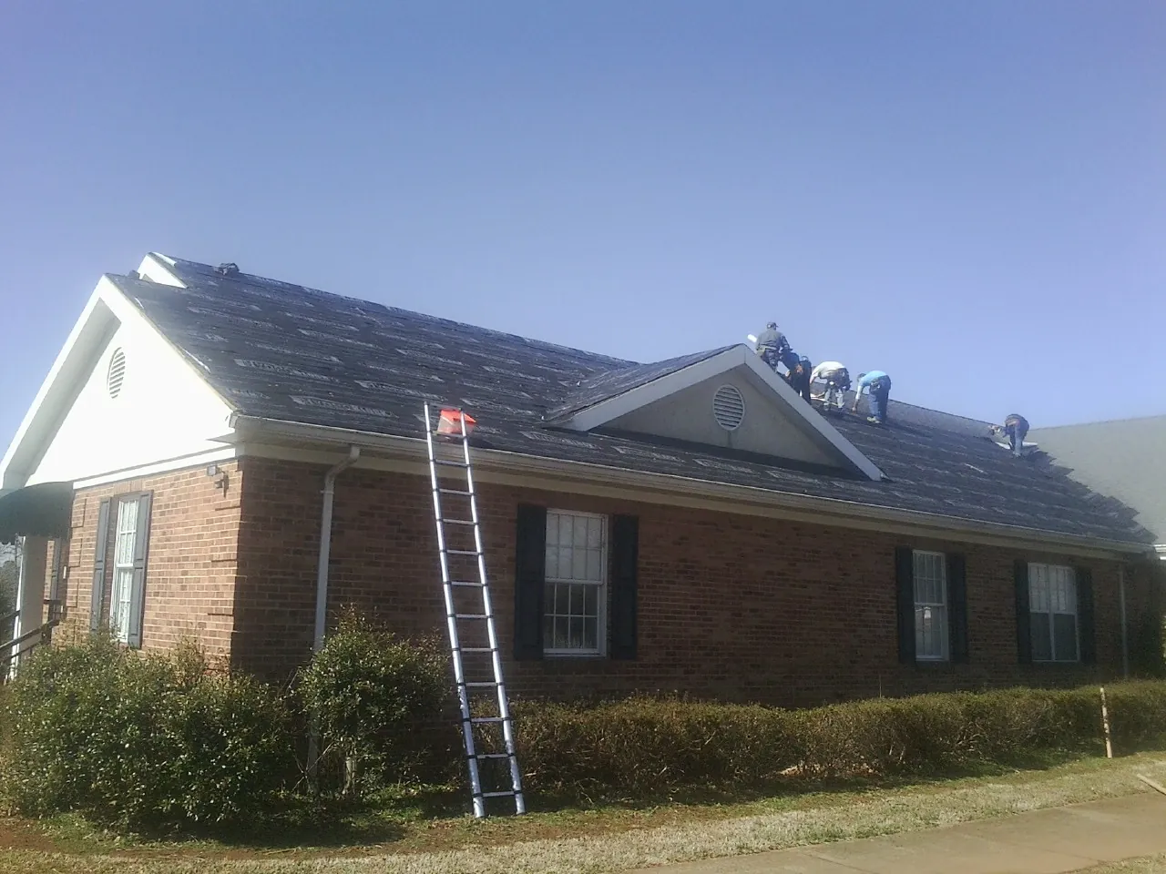Crew tearing shingles off a brick ranch roof with a ladder set — Emergency Roof Repair work by our crew in Evart