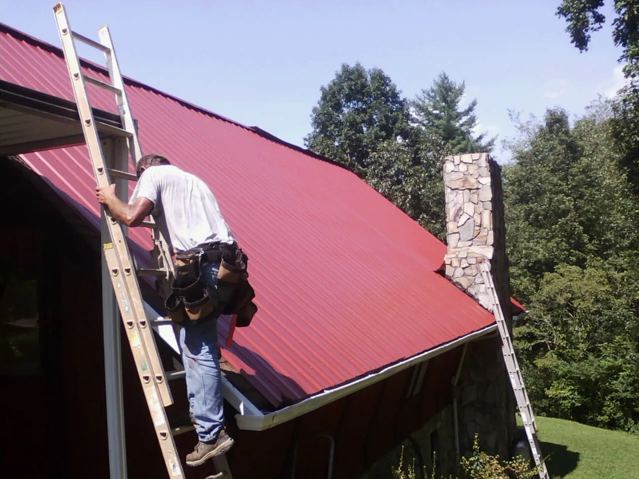 Roofer on a ladder beside a red metal roof and stone chimney — Soffit & Fascia Repair by Roofing Brothers in Evart