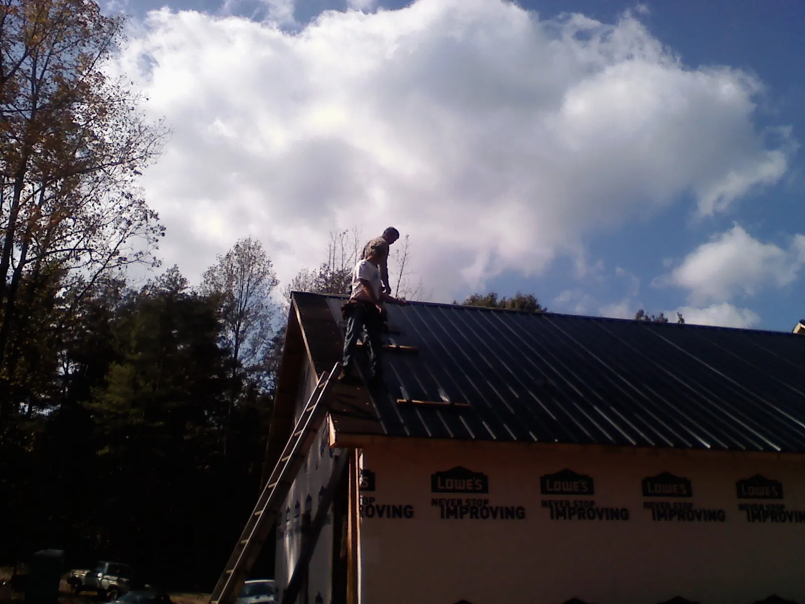 Roofer fastening dark metal panels high against a cloudy sky, from a Roof Inspection job in Evart